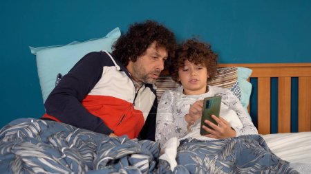 Foto de Padre e hijo niño niño 9 años de edad viendo el teléfono inteligente en línea de Internet en la cama en el dormitorio antes de seguir dormido - hábito poco saludable y la adicción a Internet en la infancia - Imagen libre de derechos