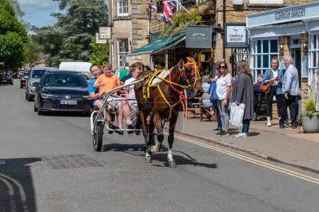 Photo for 4 young people from the Gypsy-Traveller community passing down the High Street of Bourton-on-the-Water on a 2 wheeled trotting cart pulled by a skewbald horse in bright harness - Royalty Free Image