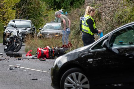 Foto de Winsley, Wiltshire, Reino Unido, 25 de septiembre de 2022, Una motocicleta gravemente dañada yaciendo en la carretera después de una colisión con un miembro del público mirando y personal del servicio de emergencia examinando la escena - Imagen libre de derechos