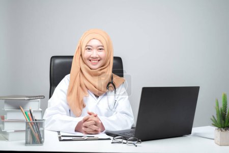 Foto de La médica musulmana asiática estaba sentada en la mesa de la paciente y miró a la cámara con una cara sonriente y acogedora.. - Imagen libre de derechos