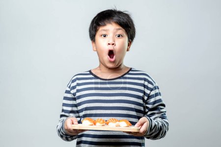 Foto de Un lindo chico asiático sostiene una bandeja de madera con sushi, salmón colocado y listo para comer con una cara feliz. - Imagen libre de derechos