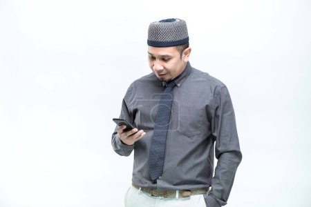 Photo for An Asian working Muslim man, dressed in an office worker, is standing and is happy to see his work done on a mobile phone with a smiling face, on white background. - Royalty Free Image