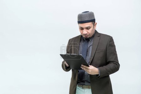 Photo for An Asian working Muslim man in an office suit is standing to check the clipboard job information with a committed face, on white background, with copy space. - Royalty Free Image