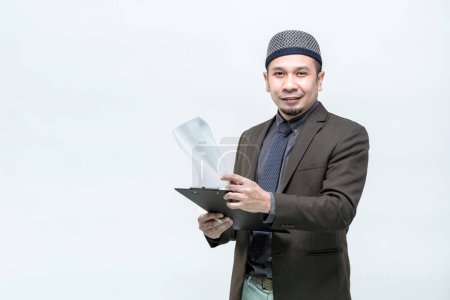 Photo for An Asian working Muslim man in an office suit is standing to check the clipboard job information with a committed face, on white background, with copy space. - Royalty Free Image