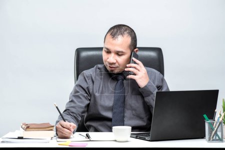 Foto de Trabajador de oficina asiático sentado frente a una computadora portátil en un escritorio y hablando en un teléfono móvil y tomando notas en la oficina. - Imagen libre de derechos