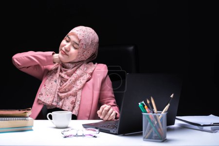 Foto de Una foto de una trabajadora de oficina, una mujer musulmana sentada horas extras trabajando en la oficina con una cara cansada y soñolienta y una taza de café en la mano. - Imagen libre de derechos