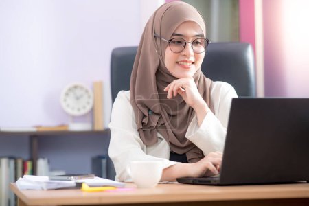 Foto de Trabajadora de oficina musulmana asiática sentada frente a la computadora portátil en el escritorio y sonriendo feliz en la oficina - Imagen libre de derechos