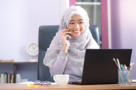 Foto de Una trabajadora de oficina para una mujer musulmana asiática sentada frente a una computadora portátil en su escritorio y hablando por teléfono móvil y trabajando en la oficina. - Imagen libre de derechos
