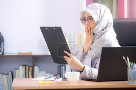 Foto de El oficinista, una mujer musulmana asiática, está sentado frente a la computadora portátil en el escritorio y está mirando los archivos con éxito en la oficina. - Imagen libre de derechos