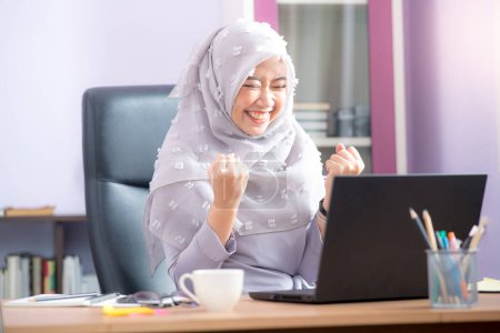 Foto de Trabajadora de oficina musulmana asiática está sentada frente a una computadora portátil en un escritorio y está muy feliz de ver el trabajo en la pantalla de la computadora en la oficina. - Imagen libre de derechos