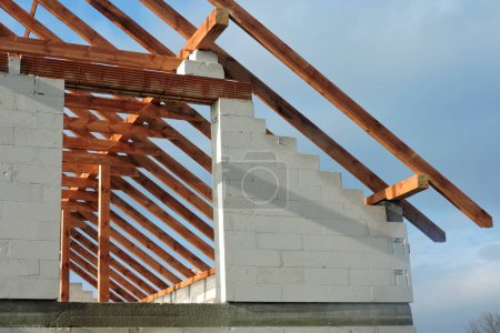Foto de Una armadura de techo de madera en una casa en construcción, paredes hechas de bloques de hormigón aireado en autoclave, una abertura de ventana áspera, un dintel de ladrillo reforzado, cielo azul en el fondo - Imagen libre de derechos
