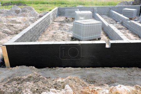 Impermeabilización de la pared de cimentación exterior con una mezcla de asfalto negro negro, un sitio de construcción con los bloques de hormigón macizo en paletas