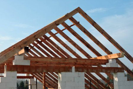 Foto de Una armadura de techo de madera en una casa en construcción, paredes hechas de bloques de hormigón aireado en autoclave, aberturas de ventanas ásperas, un dintel de ladrillo reforzado, un andamio, cielo azul en el fondo - Imagen libre de derechos