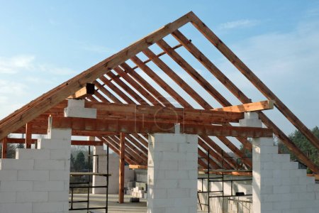Foto de Una armadura de techo de madera en una casa en construcción, paredes hechas de bloques de hormigón aireado en autoclave, una abertura áspera de la ventana, un dintel de ladrillo reforzado, un andamio, cielo azul en el fondo - Imagen libre de derechos