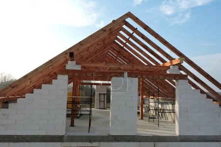 Foto de Un armazón de techo de madera de marco "A" en una casa en construcción, paredes hechas de bloques de aac, una abertura de ventana áspera, un dintel de ladrillo reforzado, un andamio, cielo azul en el fondo - Imagen libre de derechos