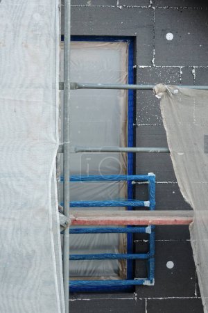 Photo for A wall of a house covered with EPS graphite polystyrene boards for thermal insulation, a scaffolding and building safety scaffolding net, a window covered with a protective film, white plastic anchors - Royalty Free Image