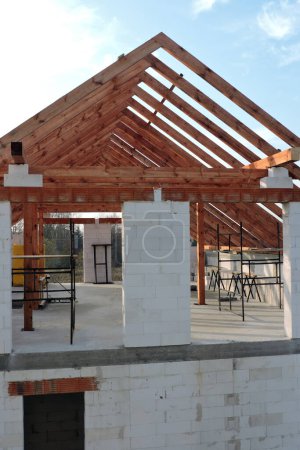 Un armazón de techo de madera de marco "A" en una casa en construcción, paredes hechas de bloques de aac, una abertura de ventana áspera, un dintel de ladrillo reforzado, un andamio, cielo azul en el fondo