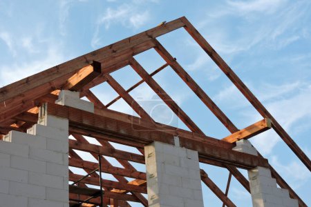 Una armadura de techo de madera en una casa en construcción, paredes hechas de bloques de hormigón aireado en autoclave, aberturas de ventanas ásperas, un dintel de ladrillo reforzado, un andamio, cielo azul en el fondo