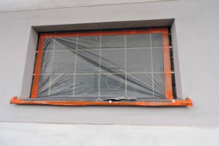 Ein Fenster aus Glasstein und eine Fensterbank, die mit einer Schutzfolie überzogen ist, eine Außenwand, die mit Grundierung bedeckt und teilweise verputzt ist