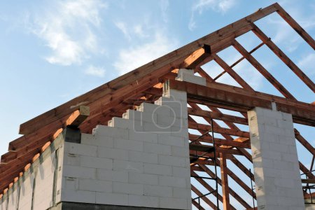 Photo for A timber roof truss in a house under construction, walls made of autoclaved aerated concrete blocks, rough window openings, a reinforced brick lintel, a scaffolding, blue sky in the background - Royalty Free Image