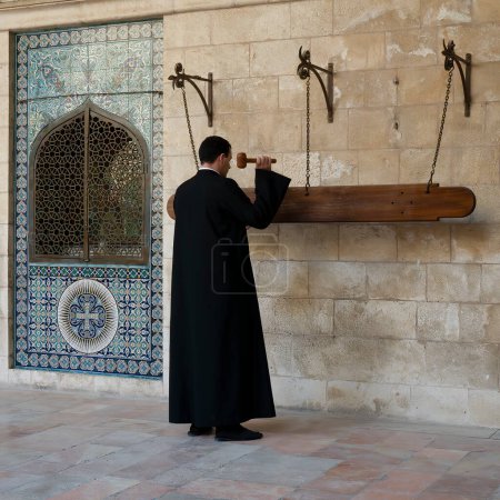 Foto de Jerusalén, Israel - 12 de noviembre de 2022: Un sacerdote armenio usando un semantrón para convocar a los adoradores a la oración en la entrada de la catedral de Santiago en el barrio armenio, Jerusalén, Israel. - Imagen libre de derechos