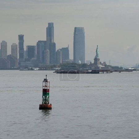 Photo for A buoy floating in the New York bay water on a hazy day, with Manhattan and the statue of liberty in the background. - Royalty Free Image