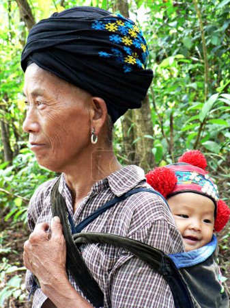 Foto de Acercamiento de la mujer Yao en sombrero tradicional con el bebé sonriente en su espalda usando brillante, sombrero hecho a mano con pompones rojos, tomada en la aldea de la tribu de la colina, Laos, Indochina, sureste de Asia - Imagen libre de derechos