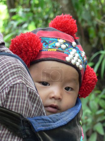 Photo for Close up of beautiful, Yao baby baby in bright, handmade hat with red pom poms, being carried on someone's back, taken in hill tribe village, Laos, Indochina, southeast Asia. High quality photo - Royalty Free Image