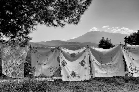 Pretty hand embroidered cloths for sale in Albania on display, hanging on washing line between trees, with beautiful mountainous scenery in background