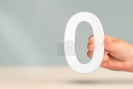 Photo for Numeral zero in hand. A hand holds a white number zero on a blurred background with copy space. Zero concept, 0 percent interest rate, minimum air emissions, cost or credit no increase - Royalty Free Image