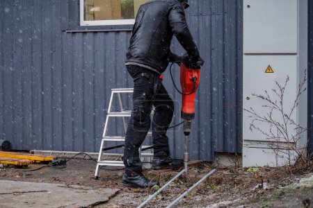 Téléchargez les photos : Un travailleur installe une tige de terre pour creuser un bâtiment. Un travailleur en vêtements de travail enfonce des tiges de terre dans le sol avec un marteau-piqueur pour éviter les courts-circuits - en image libre de droit