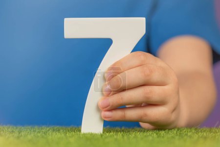 Foto de Número 7 en la mano. Una mano sostiene un número blanco siete sobre hierba verde y un fondo púrpura, siete por ciento anual. Día Mundial de la Salud. 7 de abril. Concepto de vacaciones. Foto de alta calidad - Imagen libre de derechos