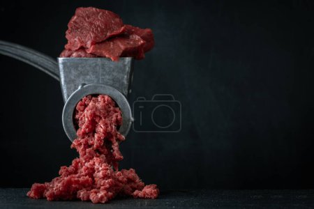 Téléchargez les photos : Fabrication de viande hachée pour boulettes de viande de bœuf dans un hachoir manuel sur fond noir. Macro shot. Gros plan de hachoir à viande métallique avec viande crue - en image libre de droit