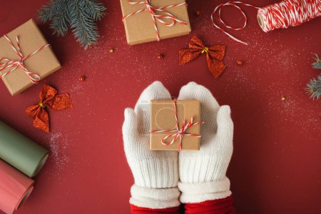 Photo for Woman hands wearing white mittens and holding cardboard gift box on the red background. Festive Christmas background with Christmas tree toys. Getting ready for upcoming holidays. Flat lay - Royalty Free Image