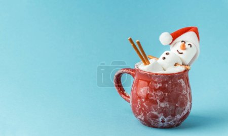 Photo for Marshmallow snowman taking hot tub in a red ceramic cup full of cocoa with milk foam. Christmas holidays blue background. Wintertime concept. Hot chocolate with marshmallow and festive decoration. - Royalty Free Image