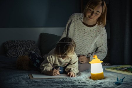 Mother and little daughter studying and drawing in a complete darkness during electricity outage. Little girl uses camping lantern to do her homework during blackout. Energy crisis concept
