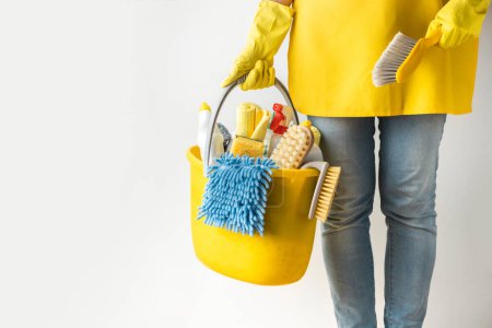 Téléchargez les photos : Unrecognizable young caucasian woman in a uniform wearing yellow rubber gloves and holding bucket full of cleaning supplies. Cleaning company service advertisement. House cleaning concept - en image libre de droit