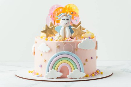 Téléchargez les photos : Unicorn cake with pink cream cheese frosting decorated with mastic rainbow, multicolored caramel candies and unicorn shaped figure on top. Birthday cake for a little girl on the white background - en image libre de droit