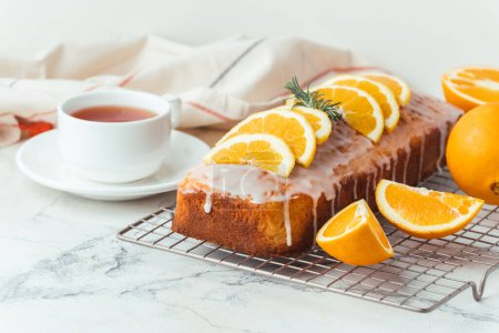 Loaf of orange bread covered with a confectionery glaze with lemon juice and decorated with orange slices. Chiffon cake on a pastry grill next to cup of tea. White background