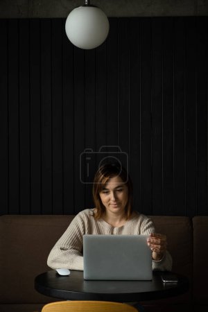 Téléchargez les photos : Belle femme caucasienne heureuse utilisant un ordinateur portable dans un espace de travail moderne. Mignon rêveuse jeune femme d'affaires assise à une table dans un restaurant - en image libre de droit
