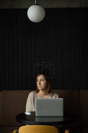 Téléchargez les photos : Belle femme caucasienne heureuse utilisant un ordinateur portable dans un espace de travail moderne. Mignon rêveuse jeune femme d'affaires assise à une table dans un restaurant - en image libre de droit