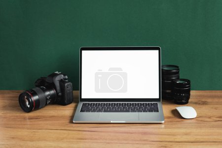Moderner Fotograf-Arbeitsplatz mit Computer, DSLR-Kamera und Objektiven. Laptop mit weißem Bildschirm auf dem hölzernen Bürotisch vor dunkelgrüner Wand. Mockup für Foto- oder Videomaker-Werbung