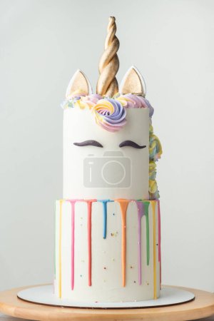 Grand gâteau superposé avec glaçage au chocolat blanc décoré de gouttes colorées, de sourcils en mastic et d'oreilles dorées avec corne sur le dessus. Fête d'anniversaire gâteau licorne pour une petite fille sur le fond blanc.