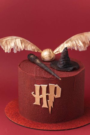 Foto de KYIV, UCRANIA - 03 de noviembre: Pastel de Harry Potter sobre fondo rojo borgoña. Tarta mágica de cumpleaños con recubrimiento de chocolate de terciopelo rojo decorado con sombrero de clasificación de masilla, varita de saúco y soplón de oro - Imagen libre de derechos