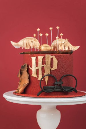Foto de KYIV, UCRANIA - 03 de marzo: Pastel de Harry Potter sobre fondo rojo borgoña. Tarta mágica de cumpleaños con recubrimiento de chocolate de terciopelo rojo decorado con lentes de masilla, varita de saúco y soplón dorado - Imagen libre de derechos