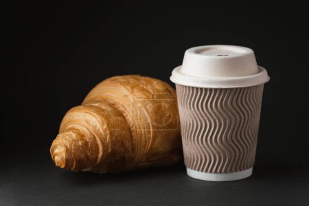 Papiertasse mit heißem Kaffee mit Papierdeckel neben frisch gebackenem Croissant auf schwarzem Hintergrund. Mock up mit Kopierplatz für einen freien Text