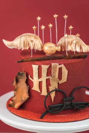 Photo for KYIV, UKRAINE - March 03: Harry Potter cake on the burgundy red background. Birthday magic cake with red velvet chocolate coating decorated with mastic glasses, Elder Wand and Golden Snitch - Royalty Free Image