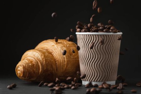Papiertasse voller Kaffeebohnen neben frisch gebackenem Croissant auf schwarzem Hintergrund. Mock up mit Kopierplatz für einen freien Text. Frühstücksroutine am Morgen