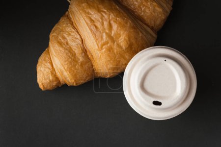 Paper cup of hot coffee with paper lid next to freshly baked croissant on the black background. Mock up with copy space for a free text. Flat lay
