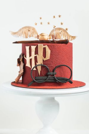 Photo for KYIV, UKRAINE - March 03: Harry Potter cake on the white background. Birthday magic cake with red velvet chocolate coating decorated with mastic glasses, Elder Wand and Golden Snitch - Royalty Free Image